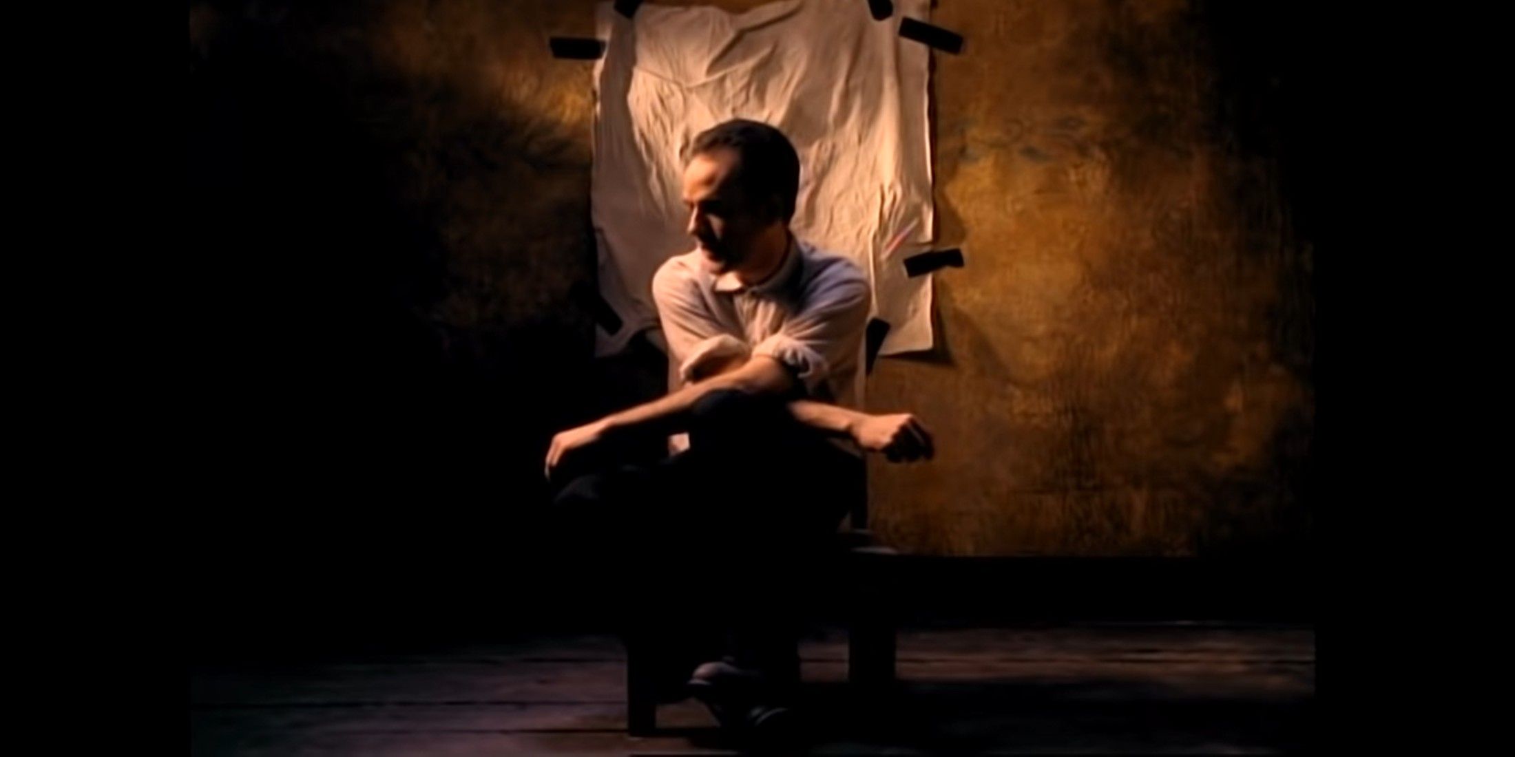 The wall in the background is brown. There is a small white sheet taped to it. In front of it, a person sits in a chair, they have a shaved head, wears a white shirt, and black pants.