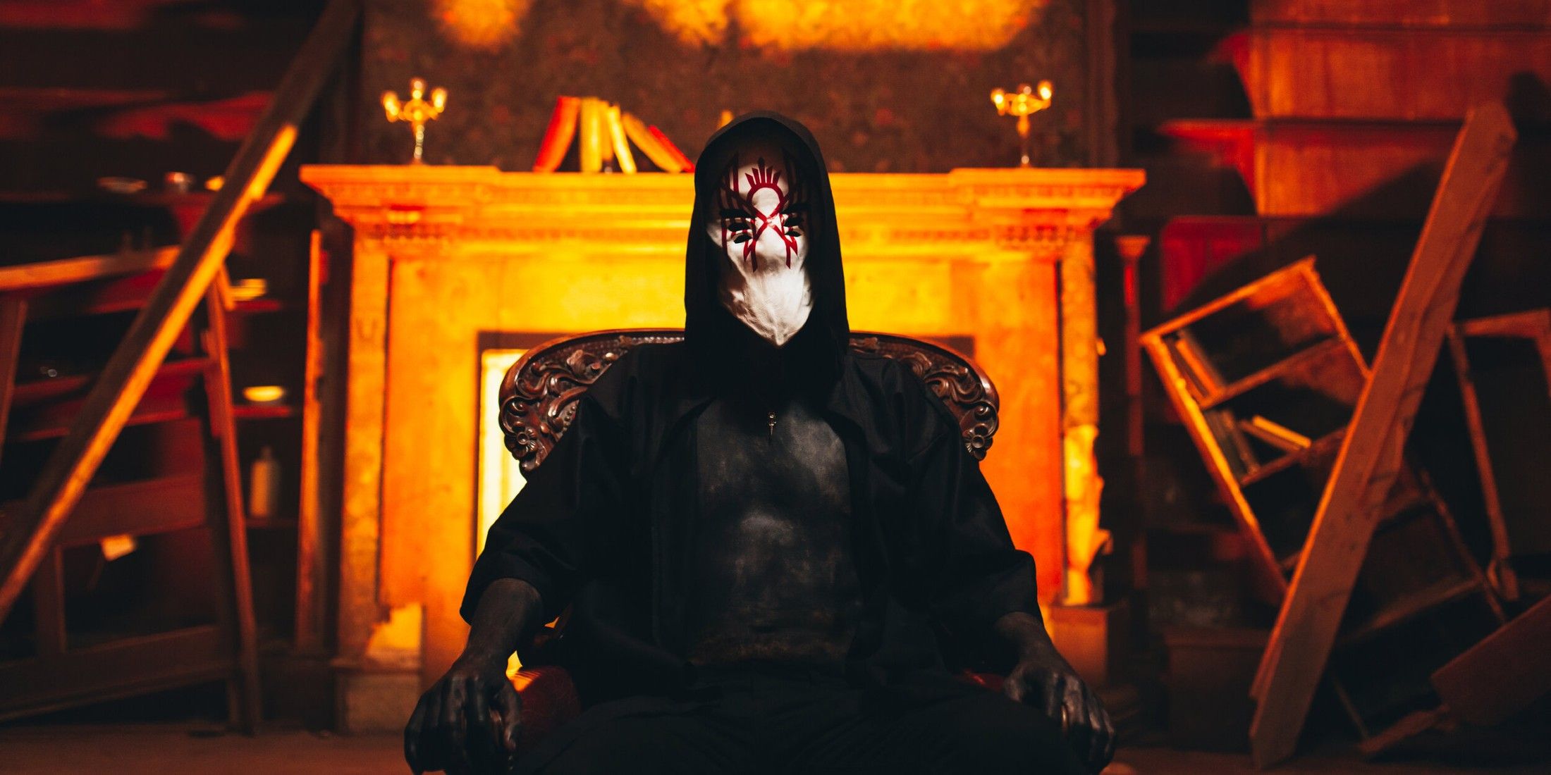 Vessel, the lead singer for the band Sleep Token, sits in a chair in front of destroyed furniture, with a fireplace illuminated by an orange glow