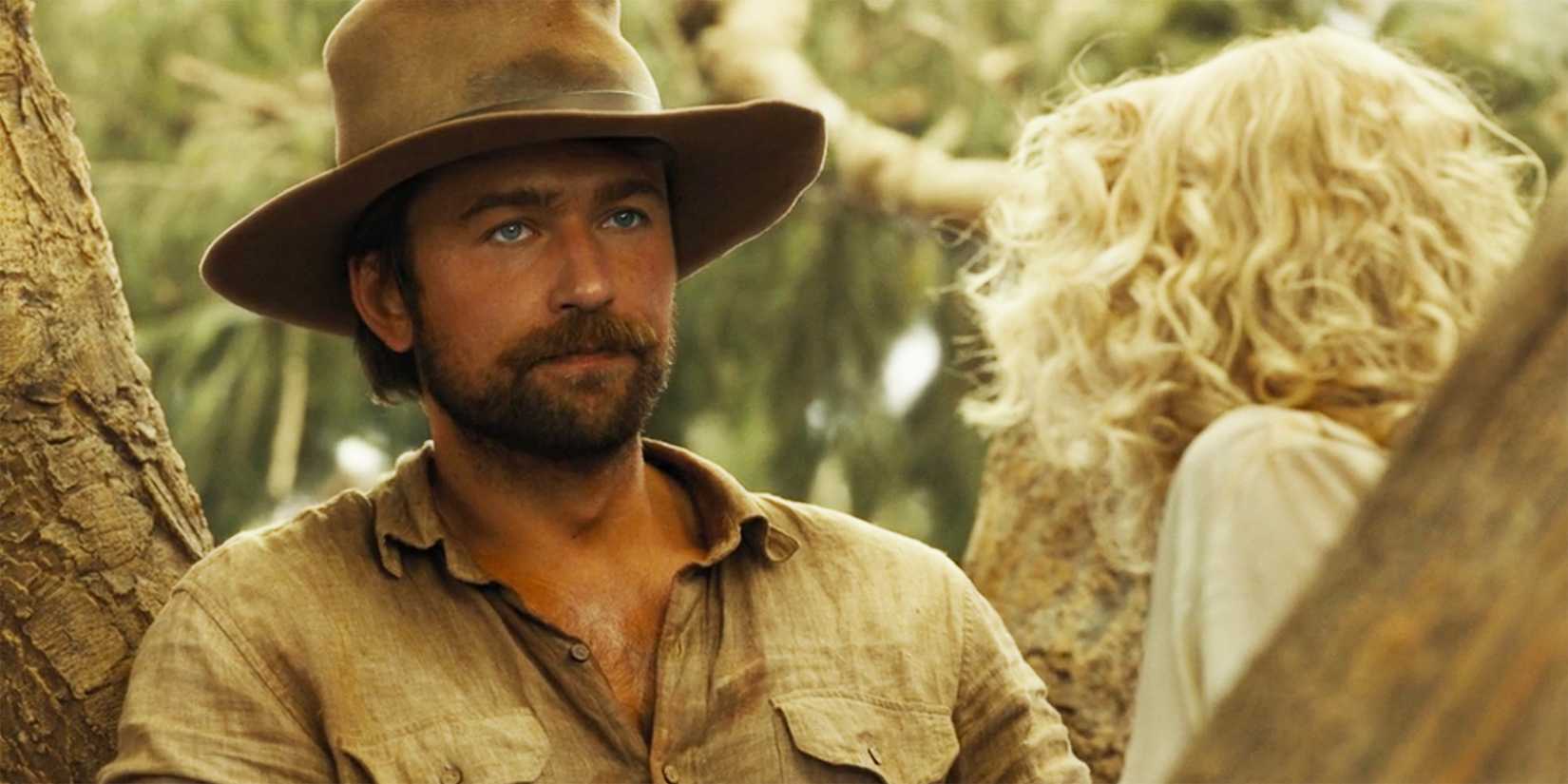 Spencer (Brandon Sklenar) sitting in a tree while wearing a cowboy hat and looking pensive in 1923