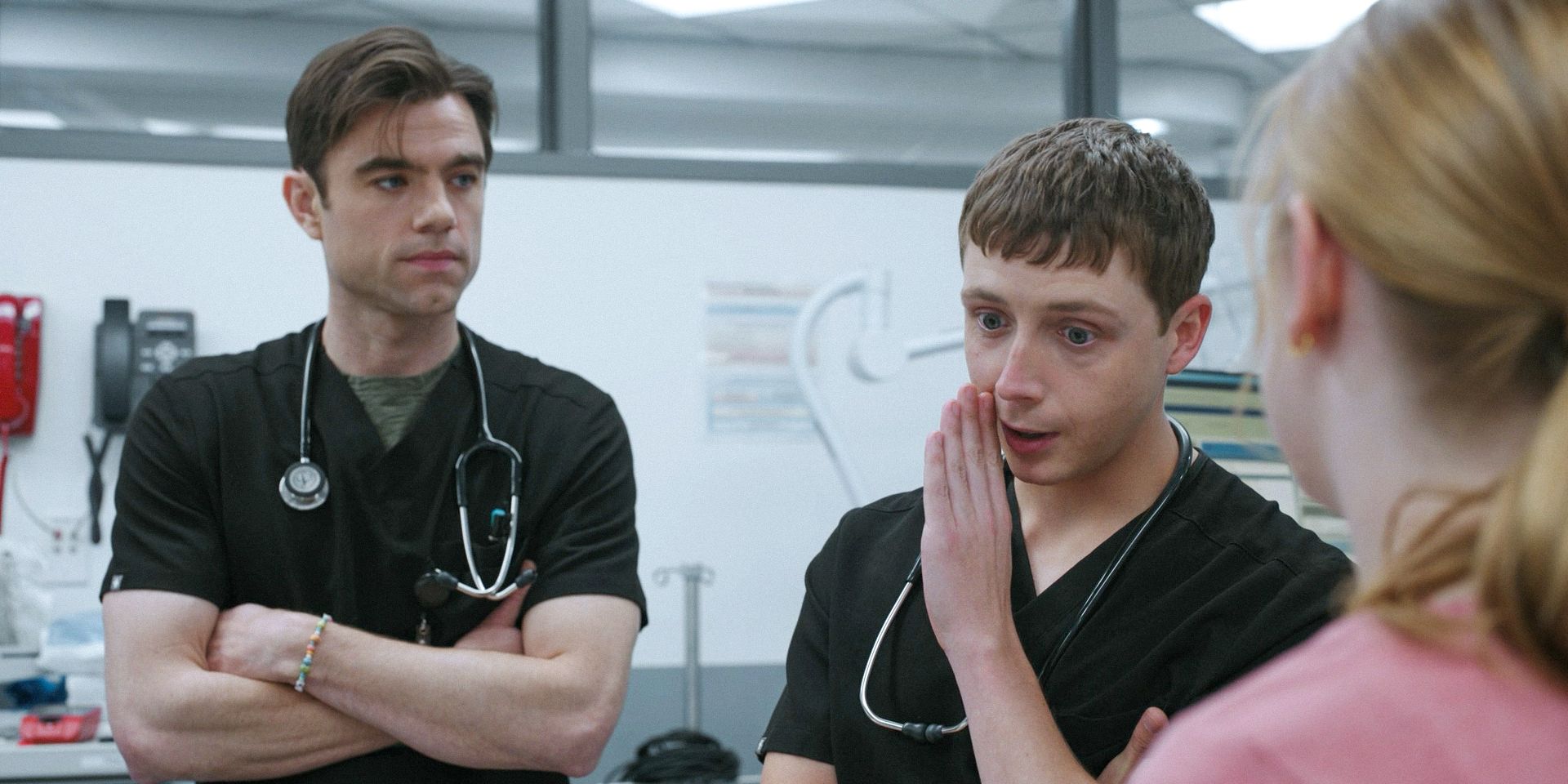 Dr. Dennis Whitaker (Gerran Howell) whispering something to a patient while Dr. Frank Langdon (Patrick Ball) looks at him with disapproval in The Pitt Season 1 Ep 10