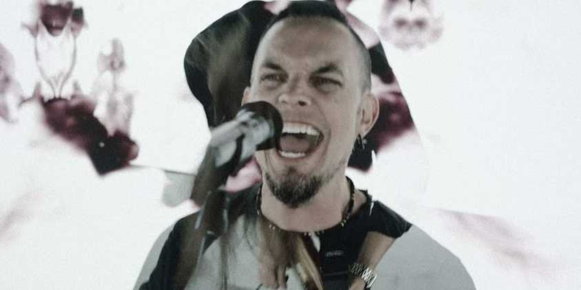 tremonti stands at a microphone with his mouth open to sing. He has a short black beard and short black hair. The background is white with splashes of roses.