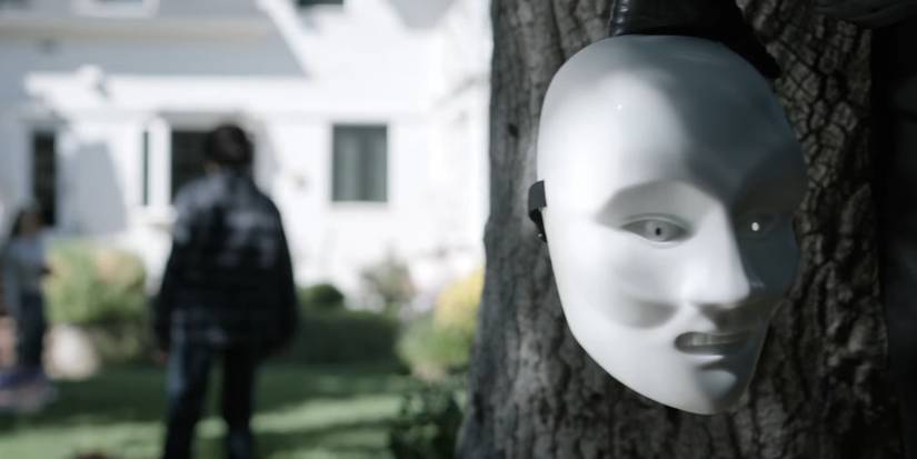A creepy white mask hanging from a tree while a shadowy figure stares at a house in Criminal Minds Evolution season 18
