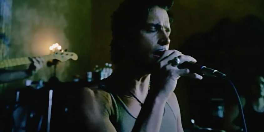 Audioslave's singer has black hair and thin mustache. He wears a grey tank top, and hold a microphone to his mouth. The wall behind him is green, and part of a bass guitar and drum are visible.