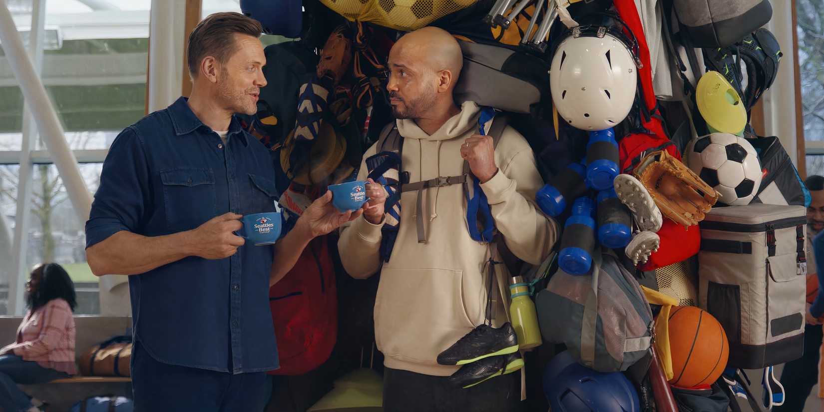 Joel McHale handing a Seattle's Best Coffee mug to a man with a large backpack full of sports equipment