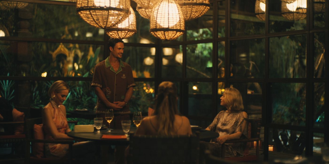 Kate (Leslie Bibb), Laurie (Carrie Coon), and Jaclyn (Michelle Monaghan) sitting at a table as Valentin (Arnas Federavicius) in The White Lotus season 3, episode 8.