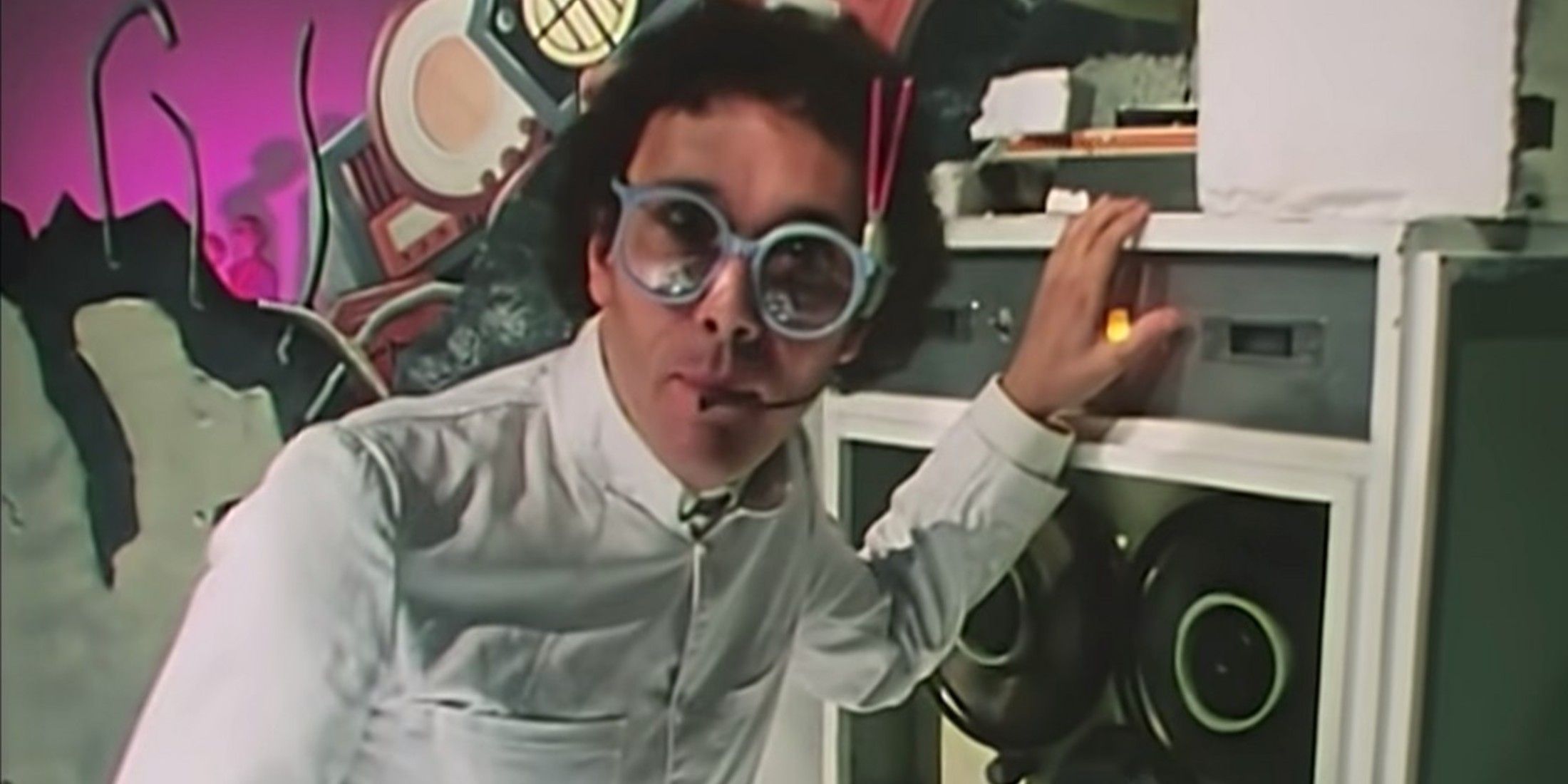 A person has on a white shirt, white glasses, with medium length hair. He stands with a hand on a radio. There is a purple painted wall with splashes of color behind him.