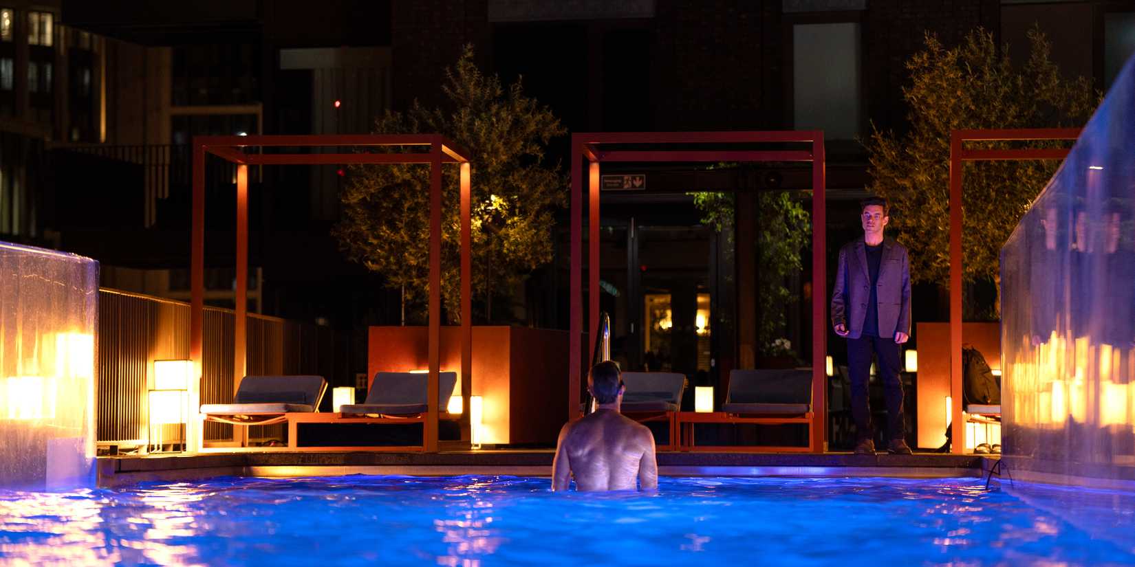 Rami Malek standing in front of a man in a pool in The Amateur