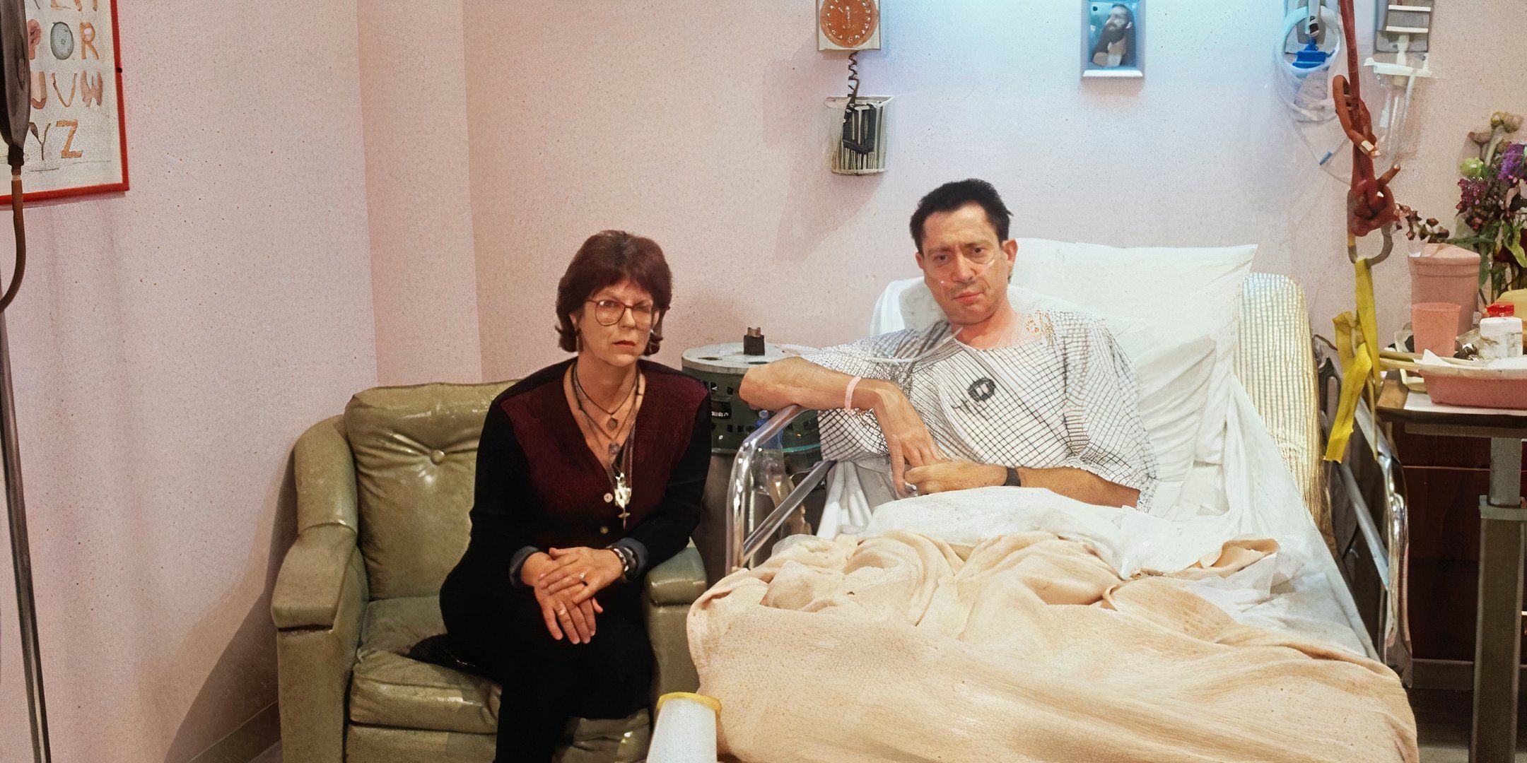 A woman looks at the camera with a serious expression as she sits next to a man in a hospital bed.