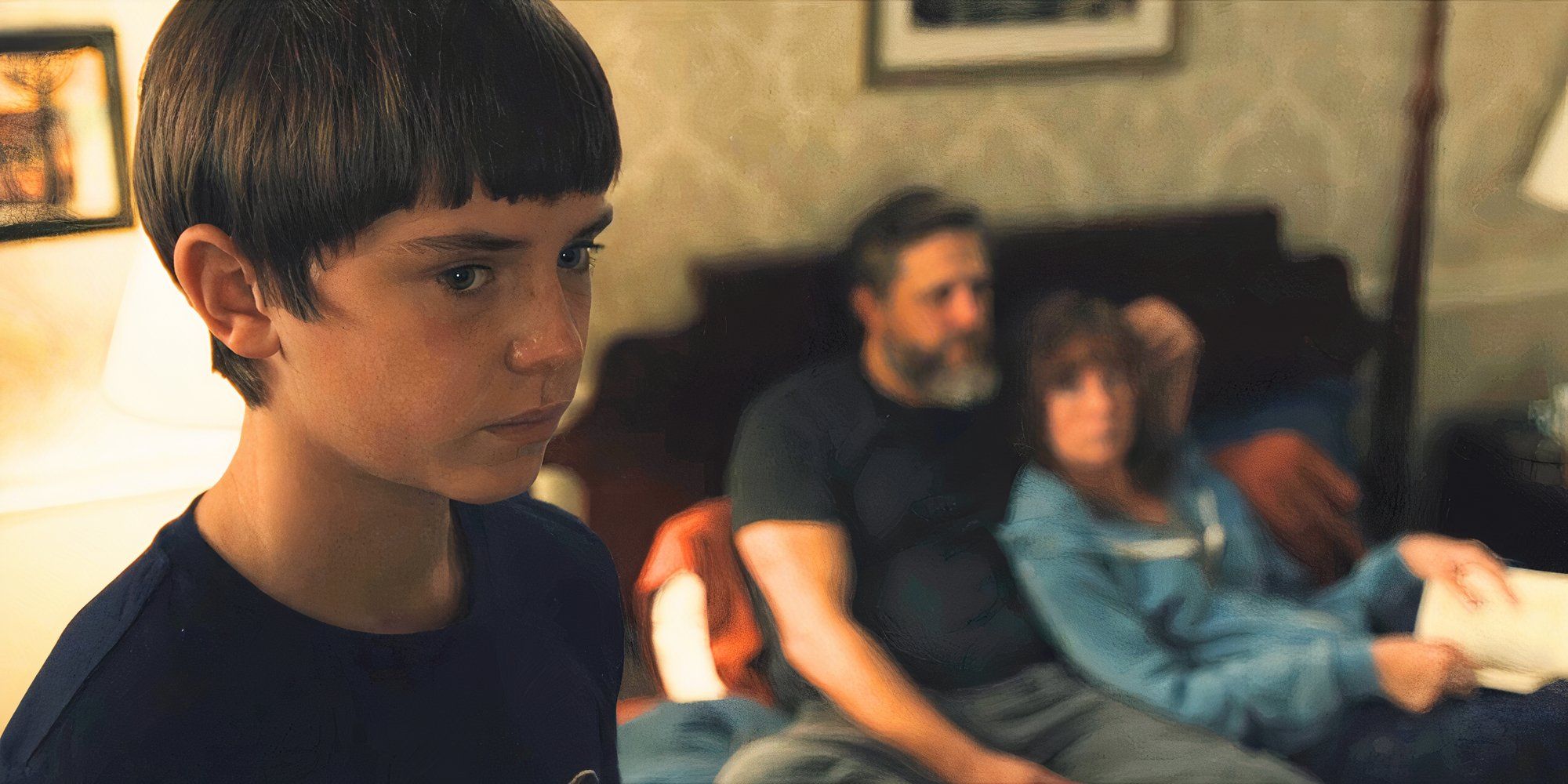 Ryan staring forward in the foreground with John and Lori on the bed in the background in Mare of Easttown