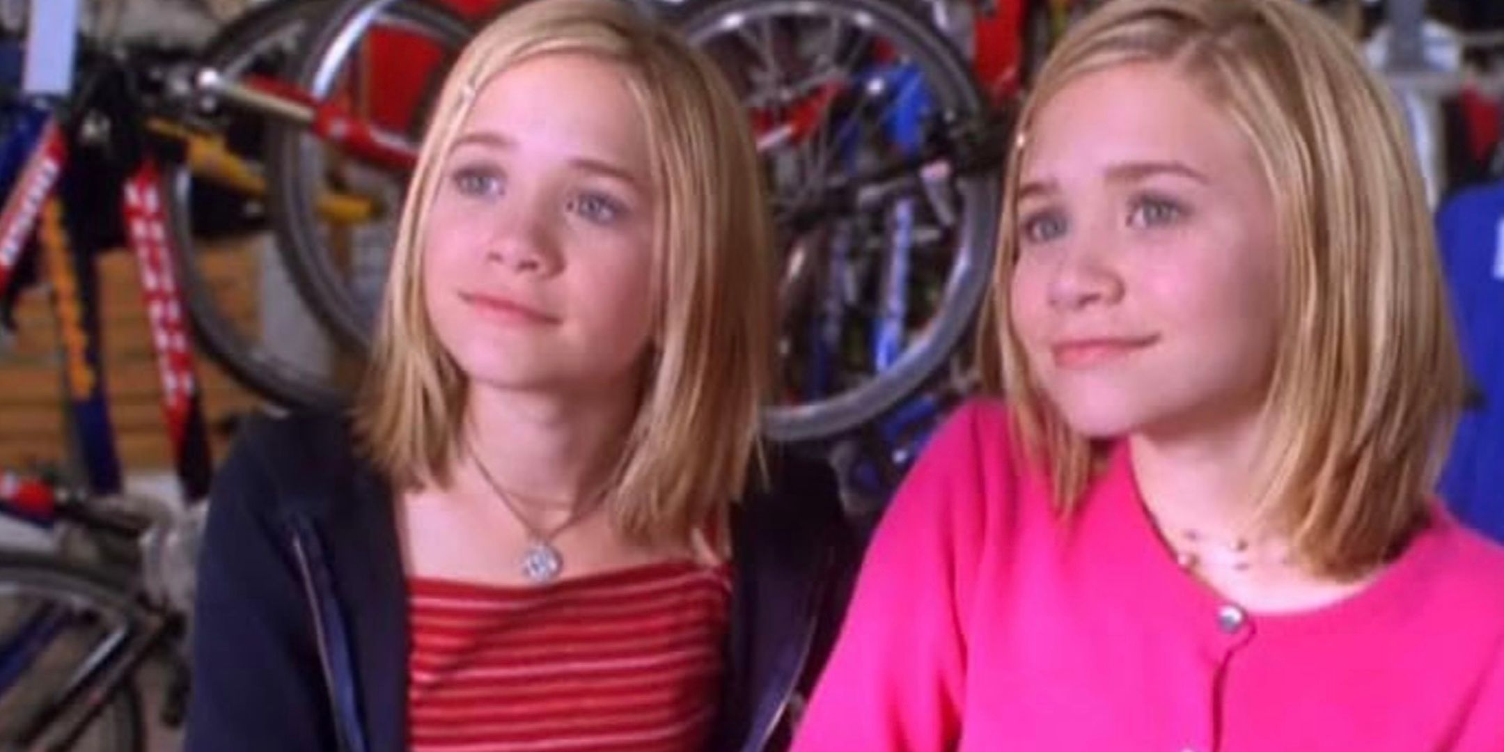 Ashley and Mary-Kate Olsen stand in front of a rack of bicycles and smile. 