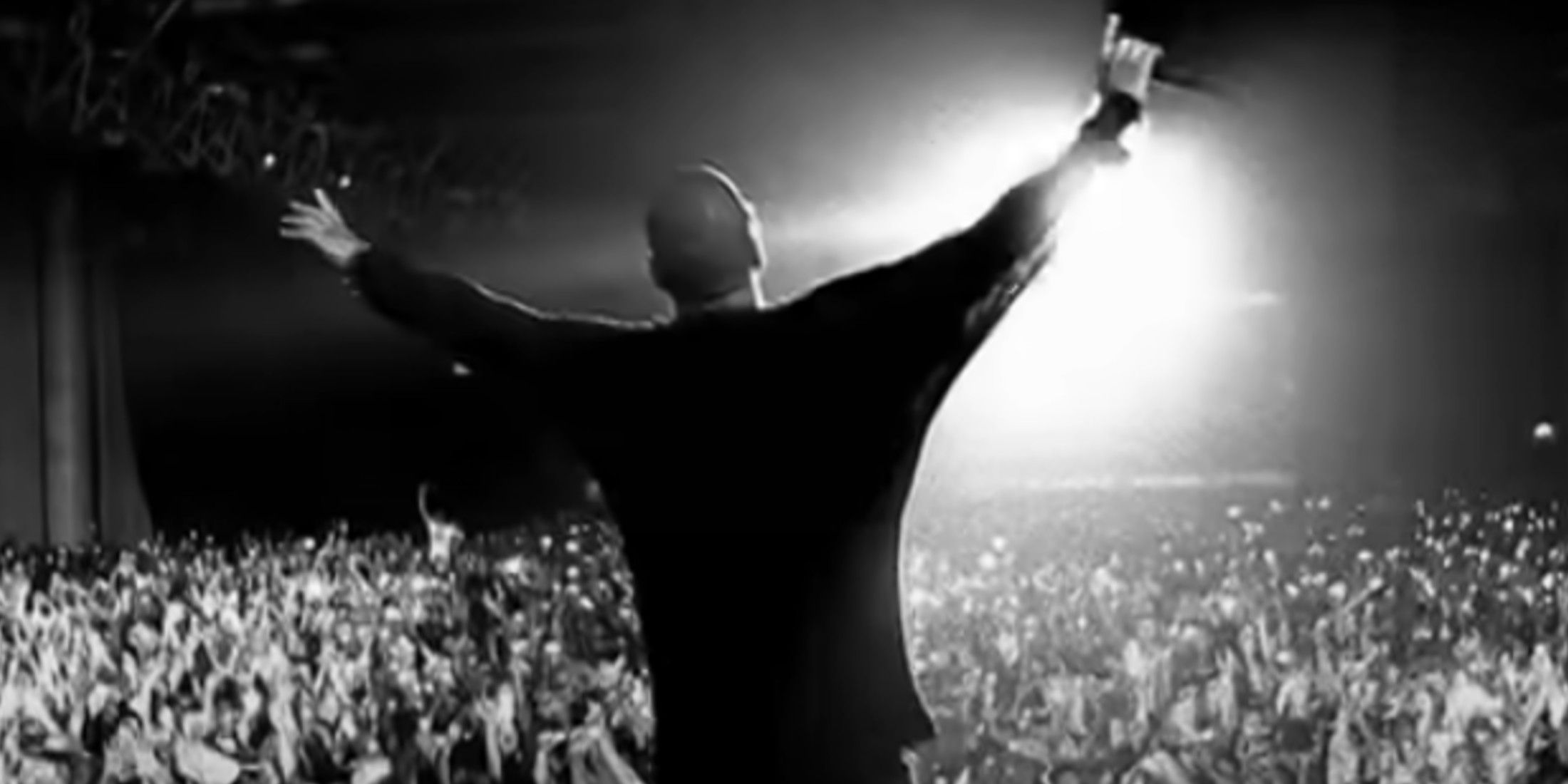 The image is in black and white. It shows Jay-Z from the waist up, holding a microphone in his left hand, with arms outstretched in front of a crowd.
