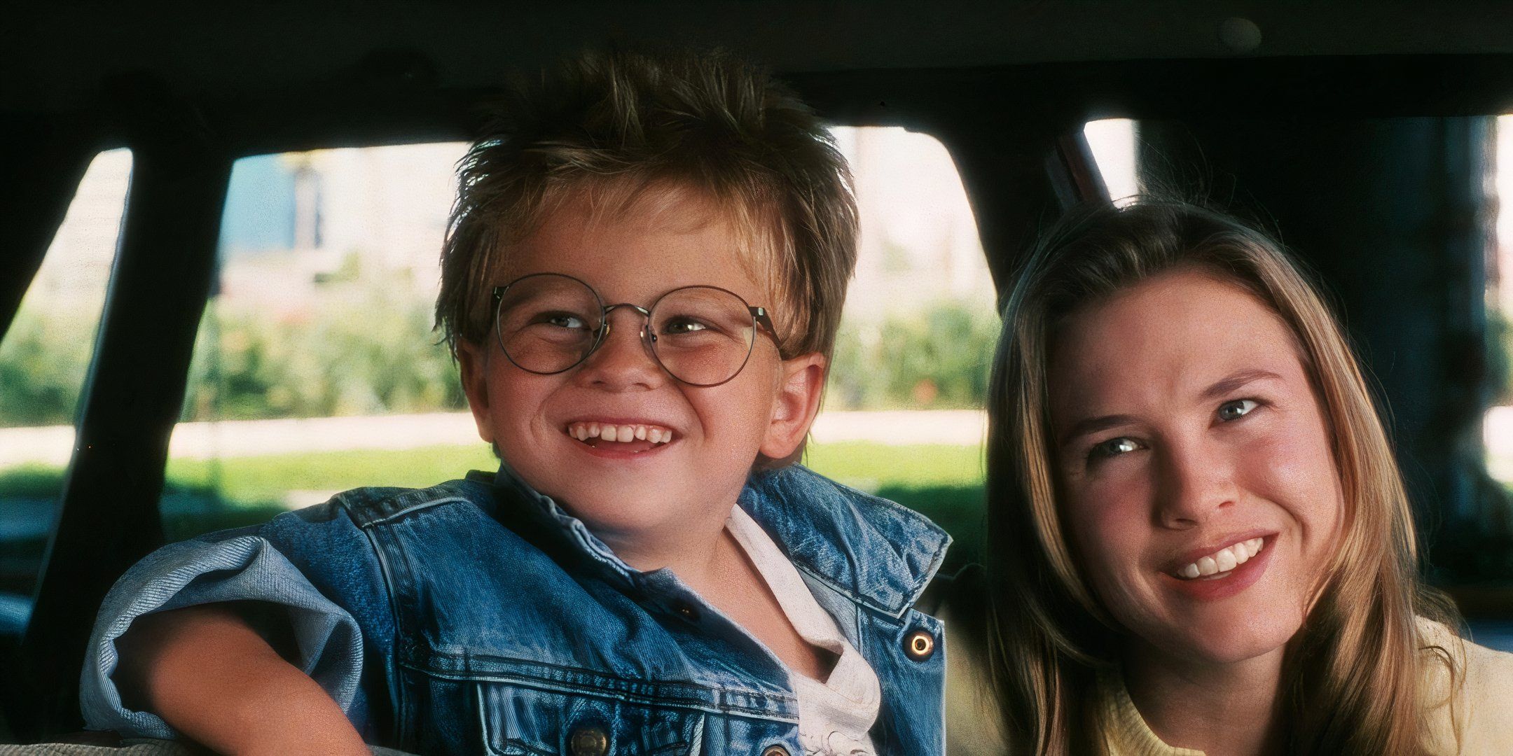 Renée Zellweger and Jonathan Lipnicki sit in the front seat of a car and smile at someone off camera.