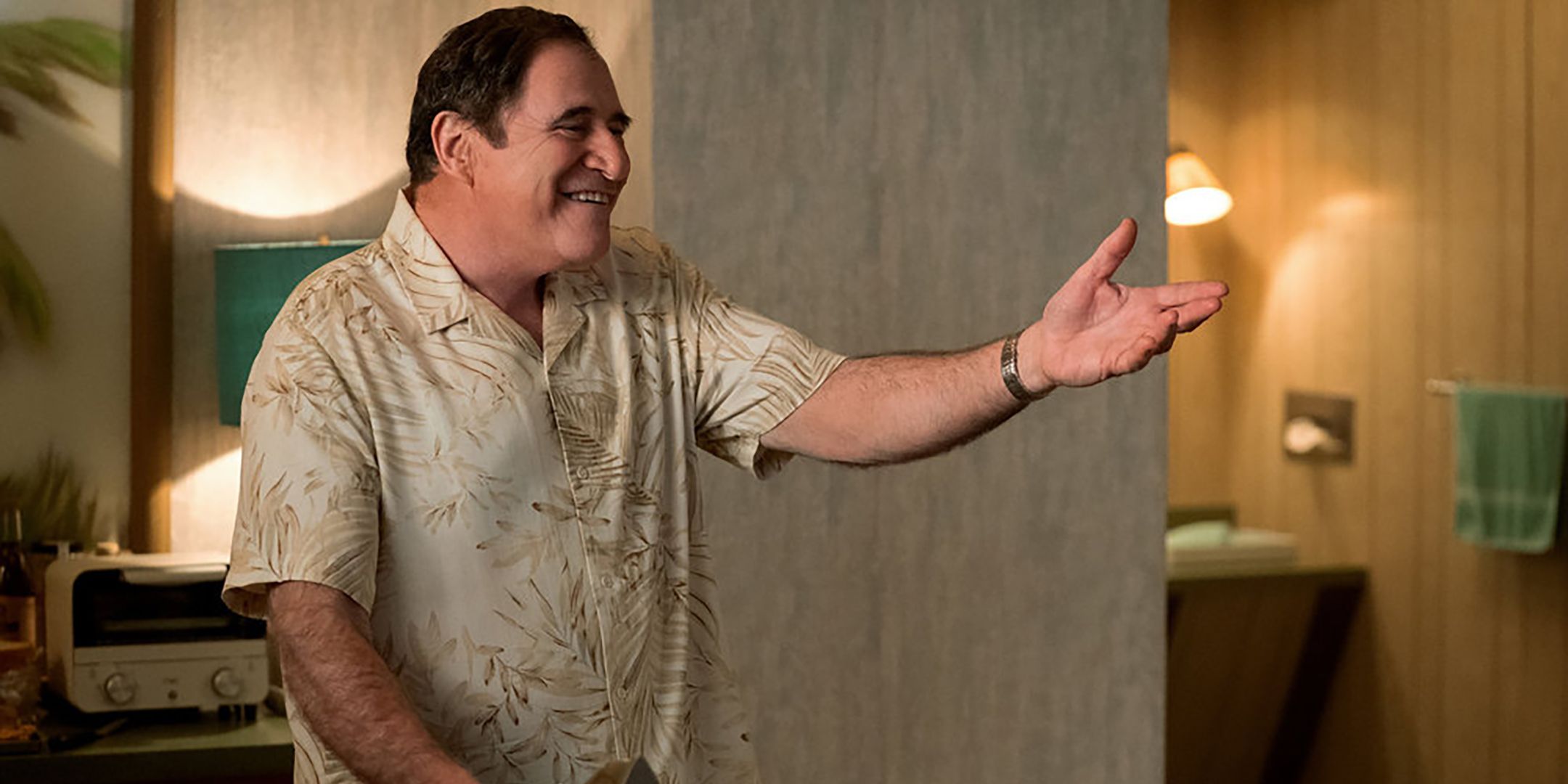 Richard Kind as Jeffrey smiling and extending his arm out in Poker Face season 2 episode 3