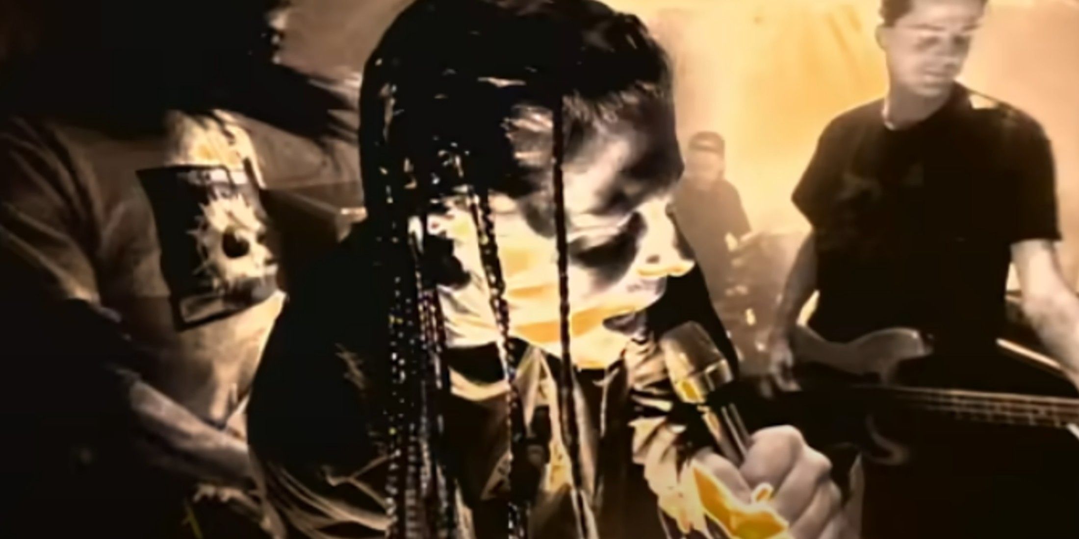 There is a yellow overlay to the photo. The guitarist is shown off to the right with his long hair, a backwards cap, and a white shirt with a black logo on it. The lead singer is shown shoulder up with his right side facing the camera. He has long braided hair, wears a long sleeve, and sings into the microphone. Behind him is the drummer who is visible to his right side. The other guitarist is on the left. He has short hair and wears a dark shirt.