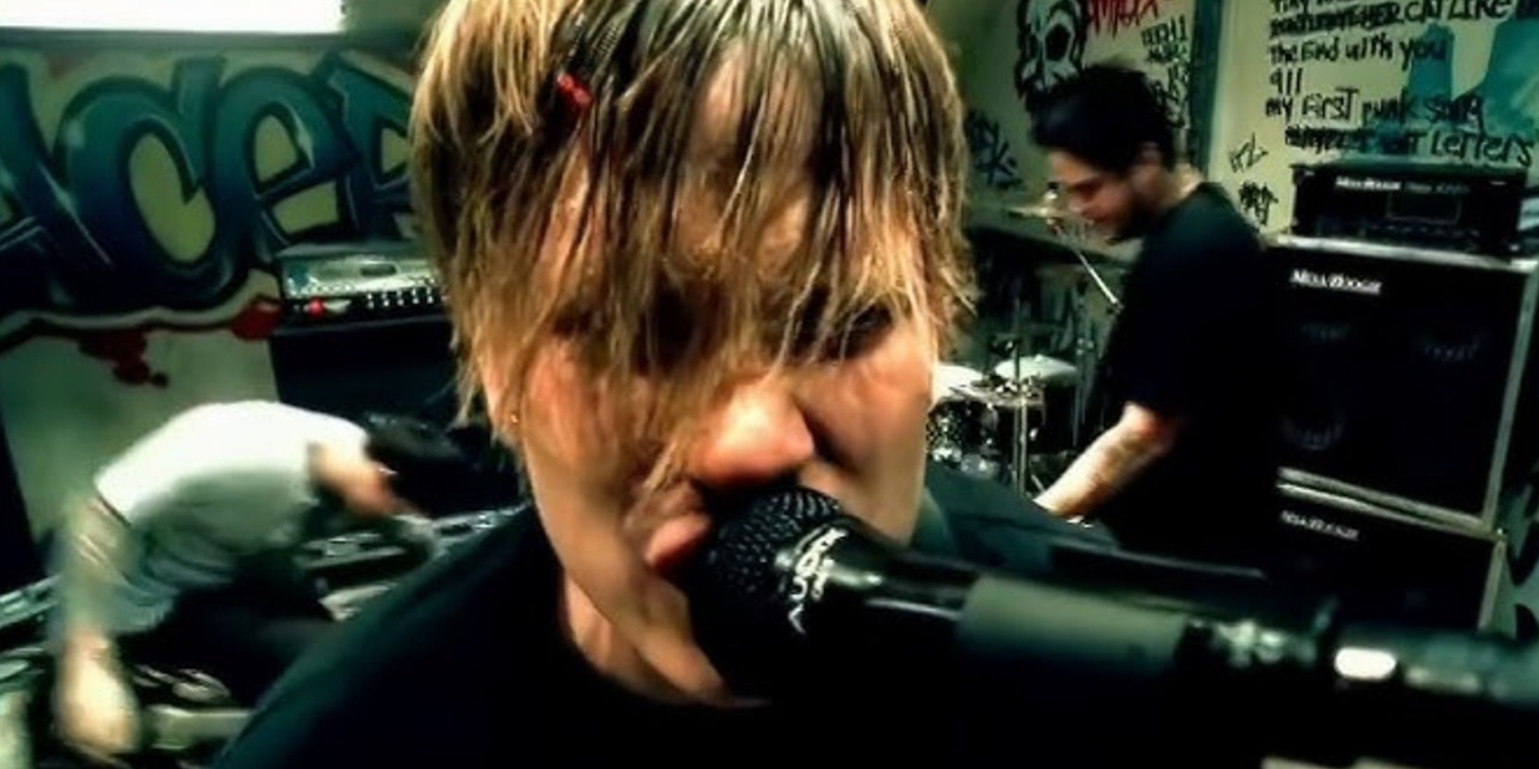 How This Beloved Drummer Went From The Local Punk Scene To Becoming A Global Icon Tom DeLonge is show from the shoulders up wearing a black shirt. He sings in a microphone with his bangs in his eyes. A guitar player is behind him in a black shirt and another guitar player is to his left in a white shirt. A small portion for an amp is visible to either side of him. There is graffiti on the back wall.