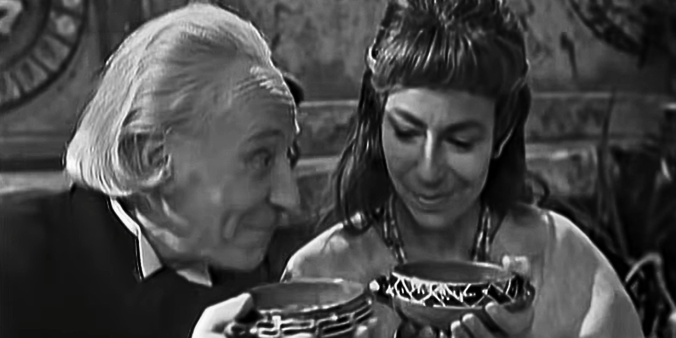 A black & white photo of William Hartnell as the First Doctor with Cameca in "The Aztecs", as the two hold up bowls
