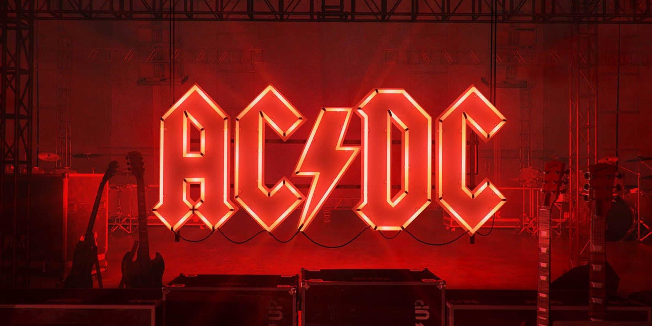 29 Years After Their Formation, Here Are The 10 Songs That Define Queens Of The Stone Age's Career The overlay of the photo is red. In the middle are the AC, followed by a lightning bolt, followed DC. A Stage apparatus is visible behind it. In front of the letters are two guitars to the left, two road cases in the middle, and two guitars to the right.