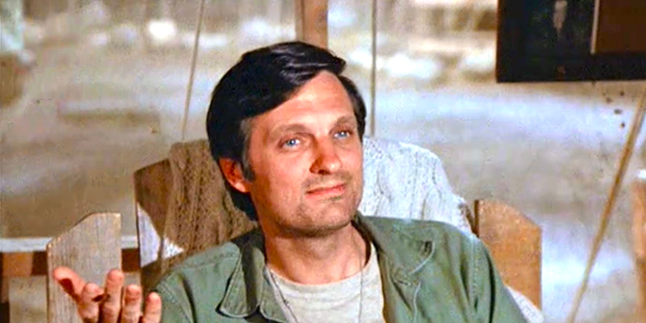 Alan Alda's Hawkeye smiles while sitting in a chair in MASH