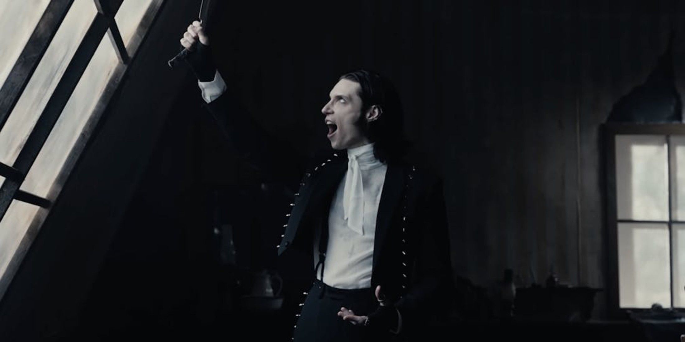 The walls in background are black. The person has on black pants, a black jacket, and a white undershirt. They also have black slicked back hair. They hold a razor to toward a window as they scream.