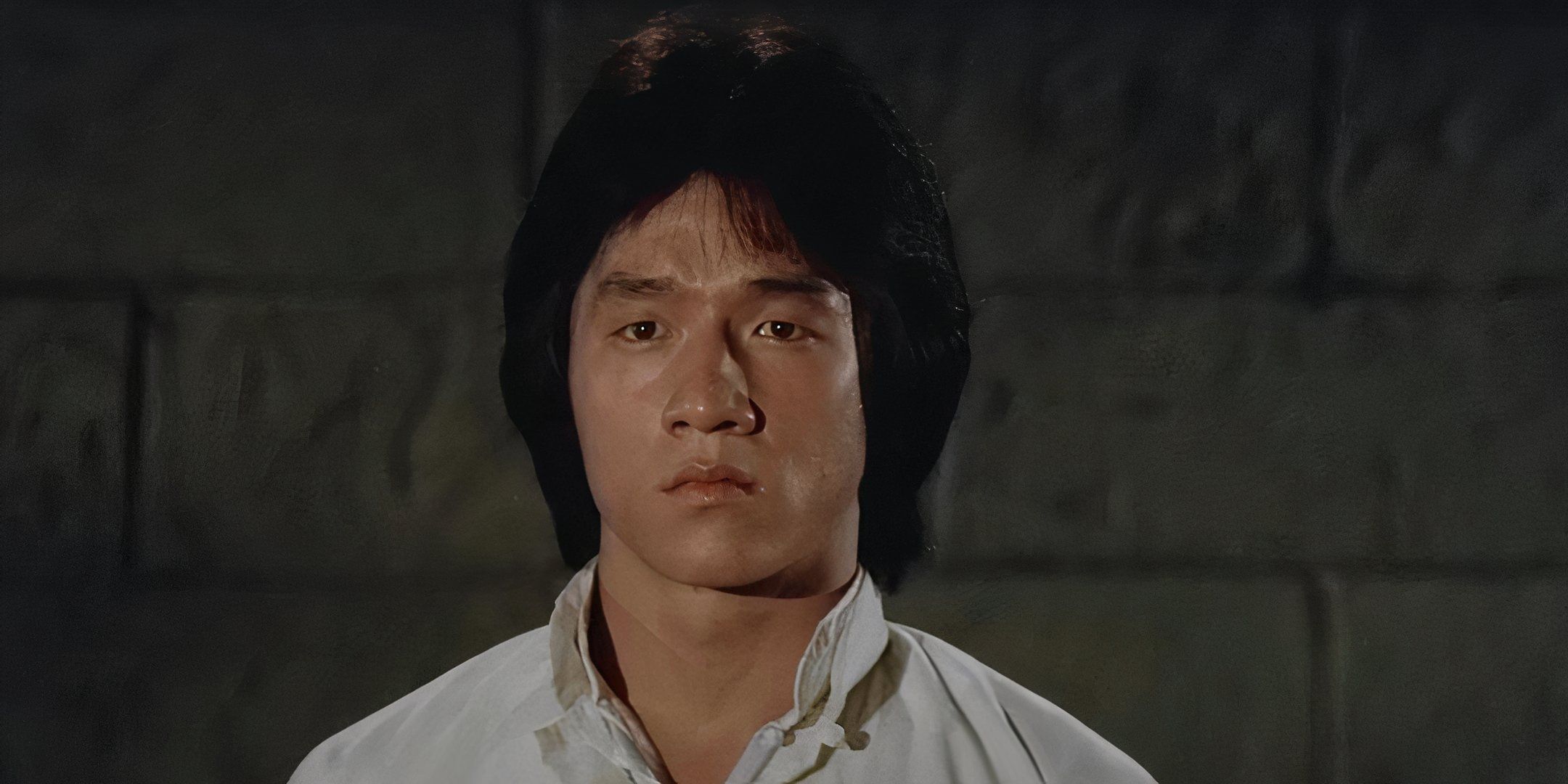 Jackie Chan is wearing a white shirt and looks ahead with a blank stare. 