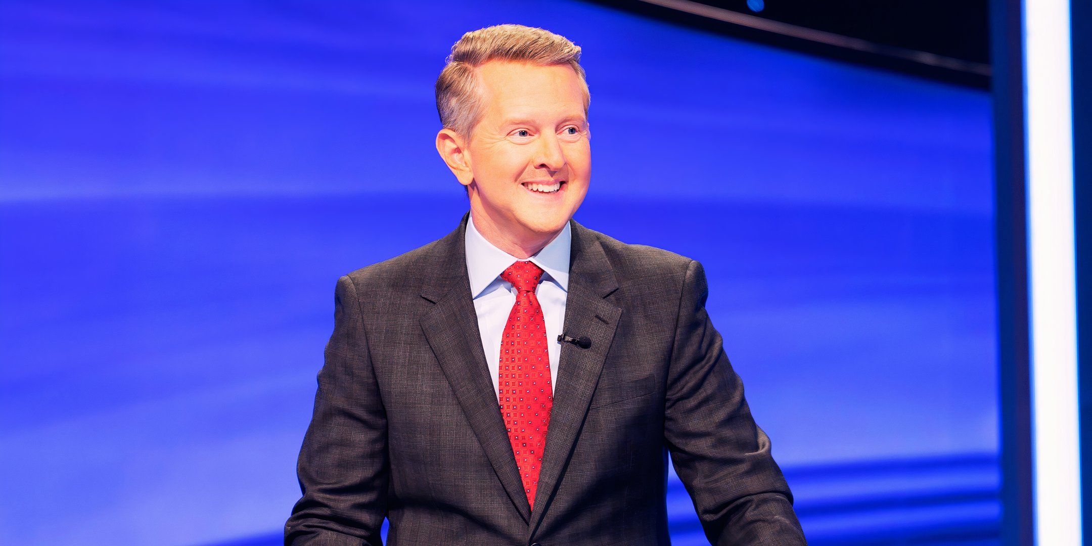 jeopardy host ken jennings smiling in a suit with a blue background