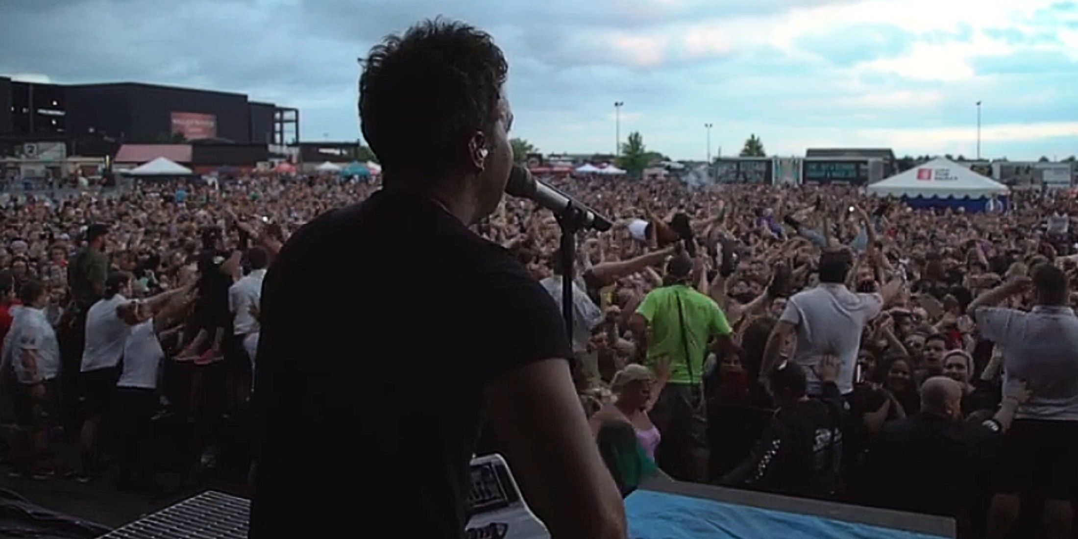 How This Beloved Drummer Went From The Local Punk Scene To Becoming A Global Icon The back of a singer on stage is visible, as he faces the crowd. He wears a black shirt, holds a guitar, and sings into the microphone. The sky is blue, with minimal clouds. There is a sea of people in front of the stage. Some pop-up tents are seen in the back.