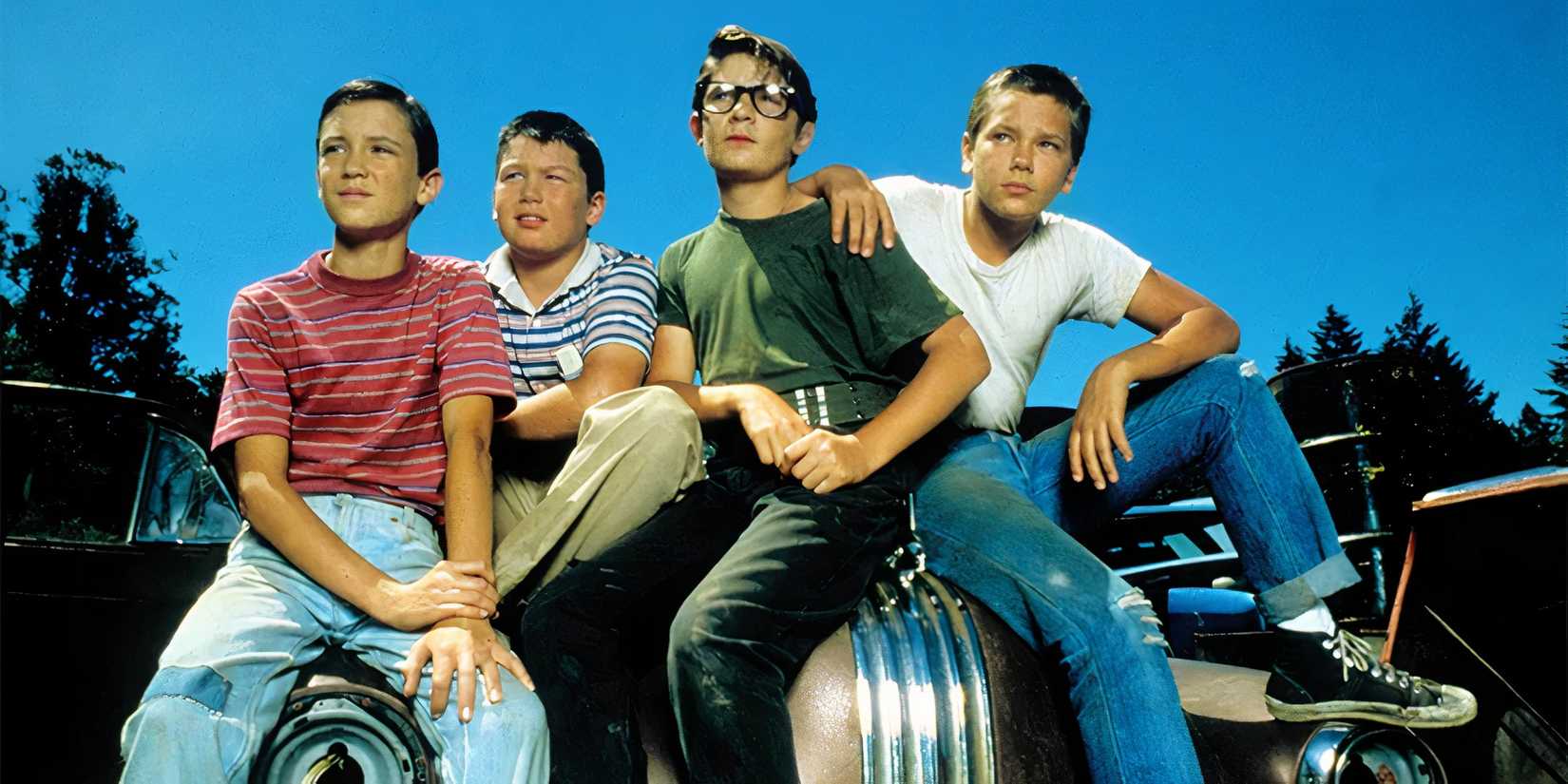 The cast of Stand By Me sit on the hood of a car looking off into the distance.