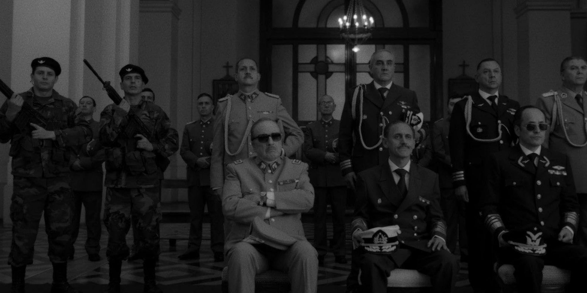 A group of armed men stand behind sitting, uniformed political leaders. 