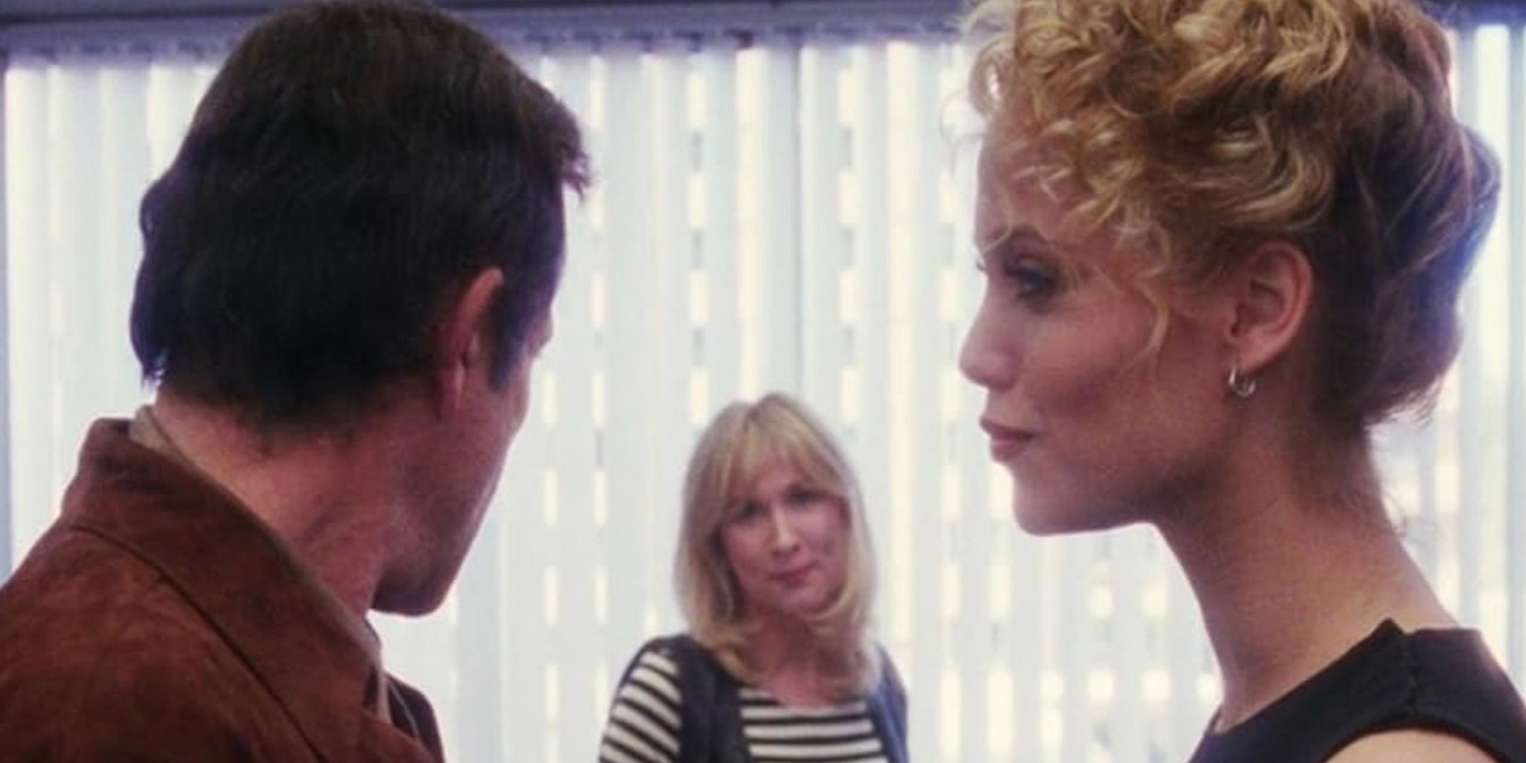 Elizabeth Berkley stands in an office, smirking while looking into the distance.