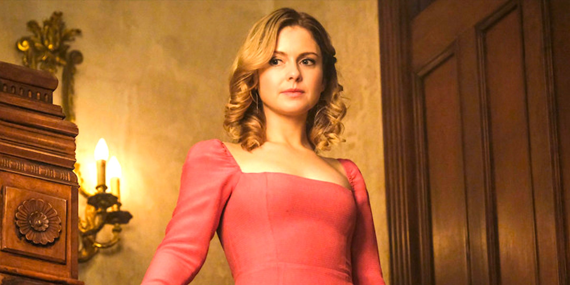 Rose McIver's Sam stands on a staircase in a pink dress from Ghosts