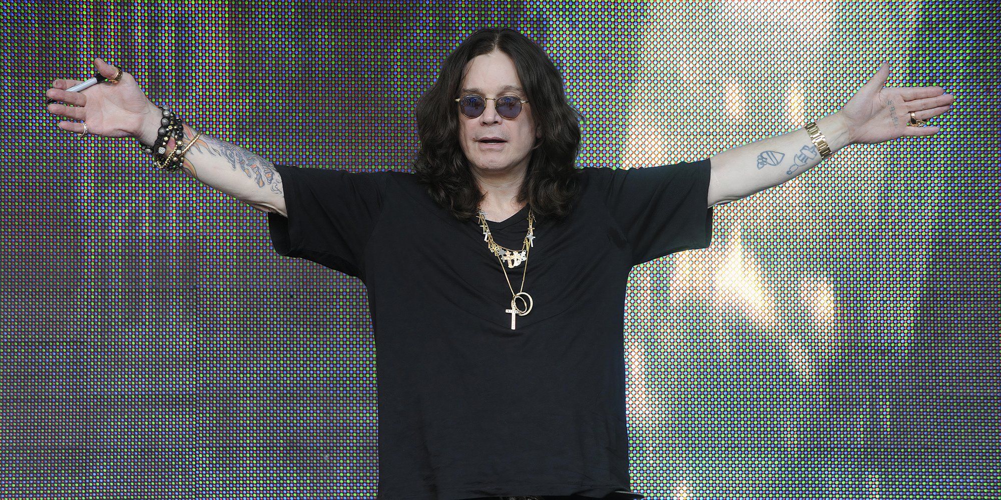 Ozzy Osbourne usa todo o preto e levanta os braços no palco enquanto usa seus óculos icônicos.