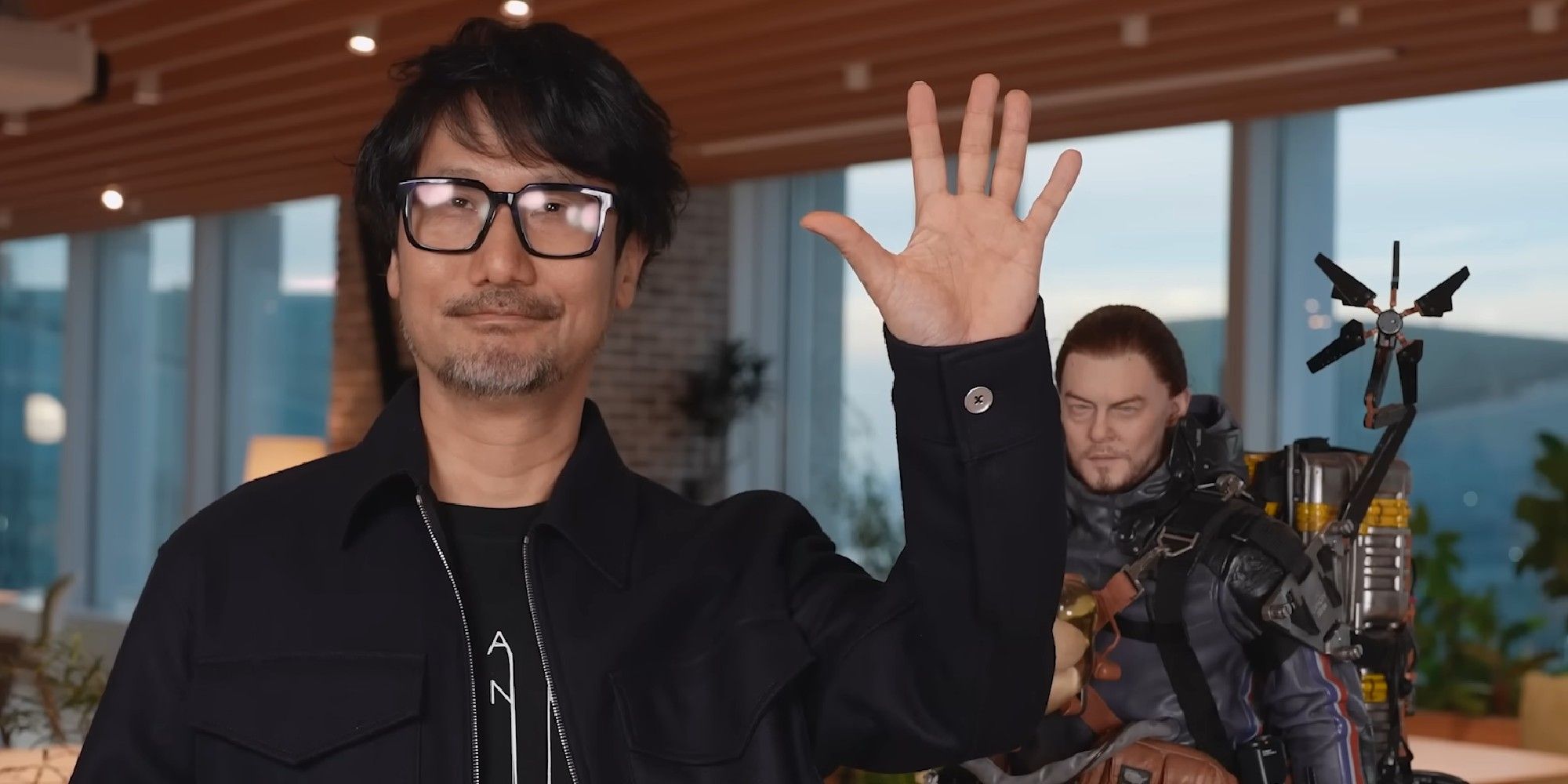 Hideo Kojima waving with statue of Sam from Death Stranding