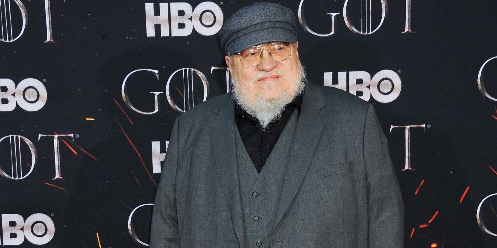 George R.R. Martin standing in front of a wall with the Game of Thrones and HBO logos on it