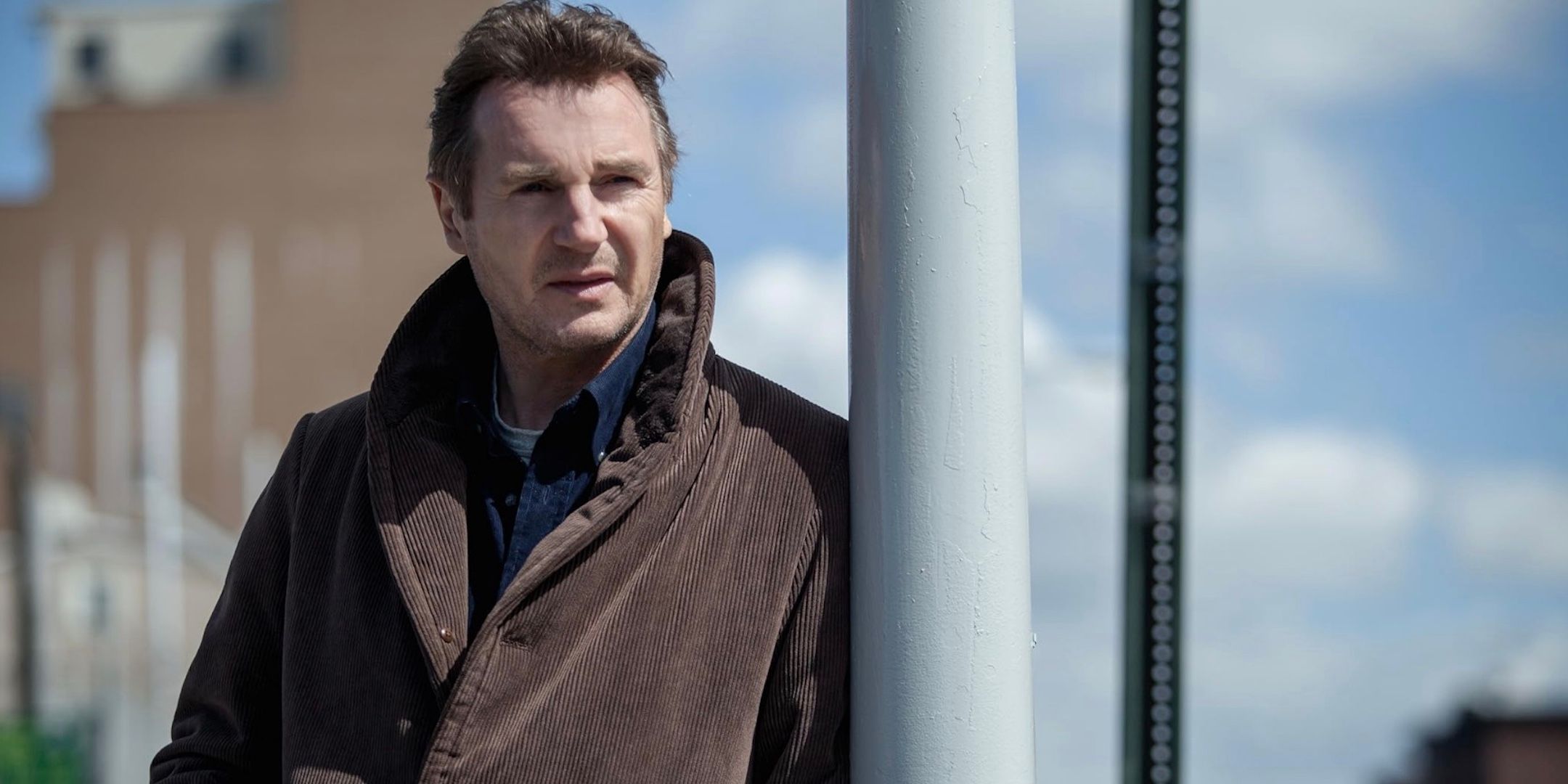 Liam Neeson is standing leaned against a metal pole. 