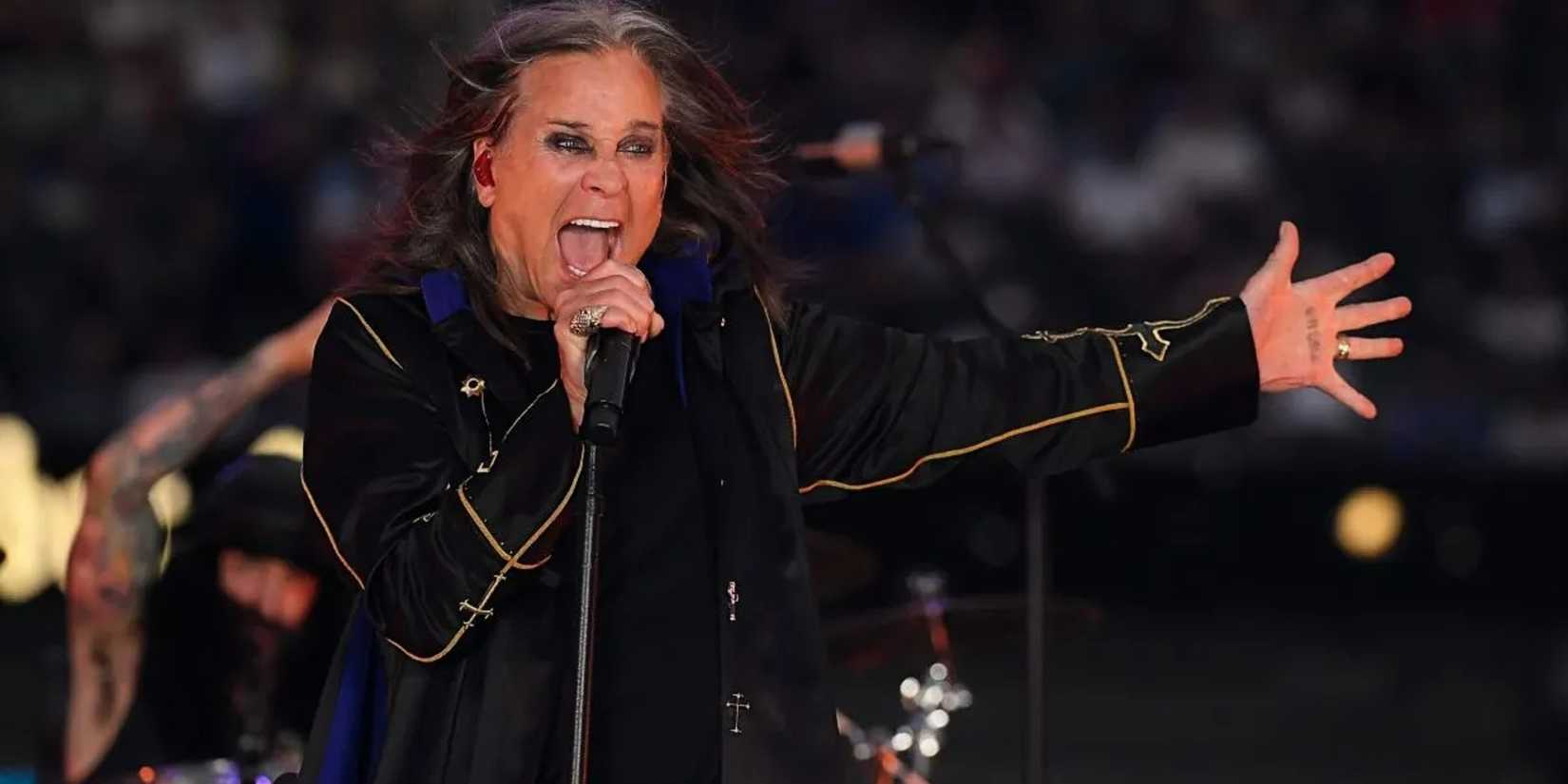 A performer passionately sings on stage, holding a microphone with one hand and extending the other. The setting conveys energy and excitement, with a blurred audience in the background.