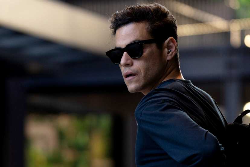 Rami Malek with sunglasses on looking to the side as he puts on a backpack in The Amateur