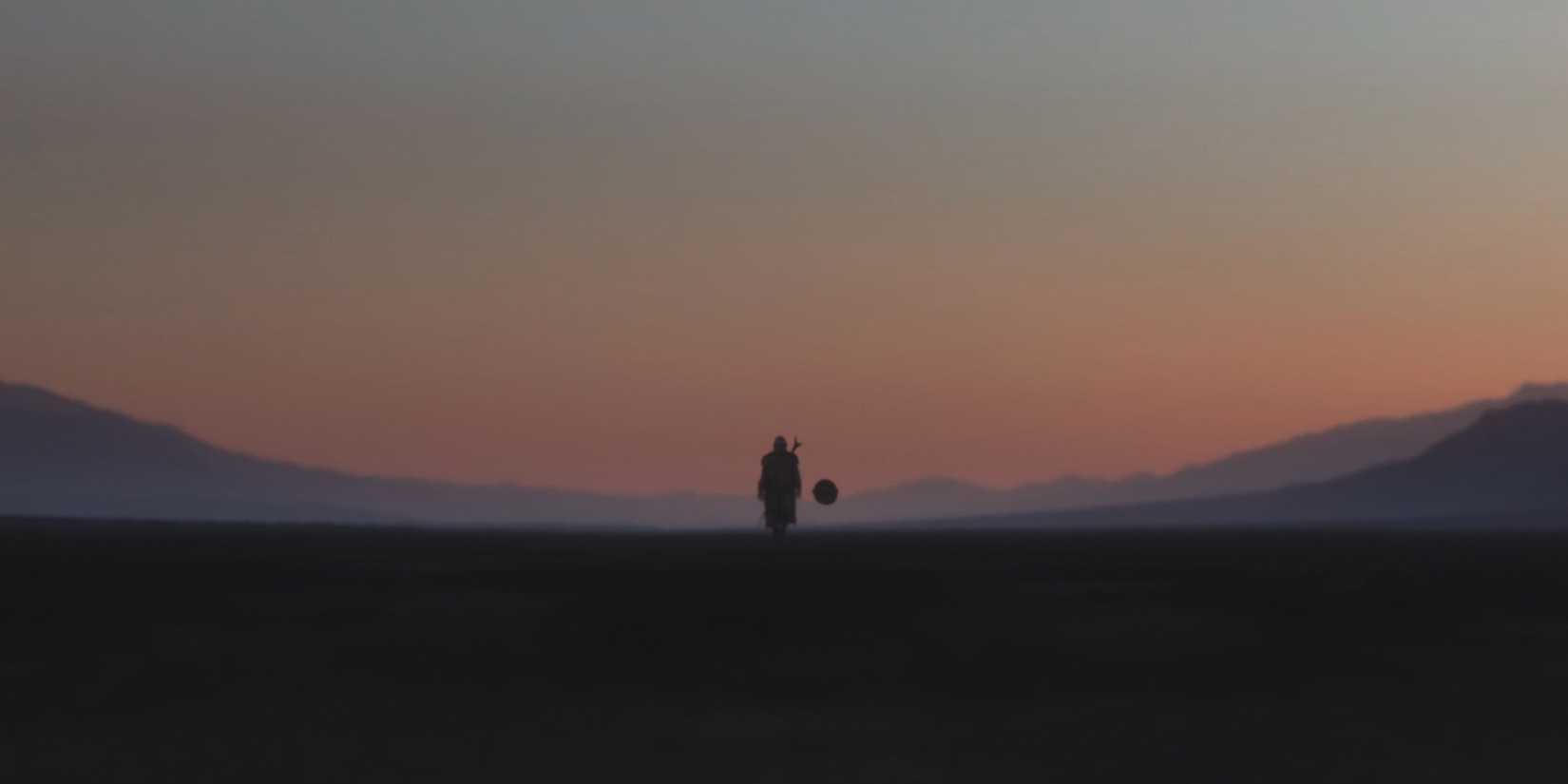 Din Djarin walks alongside Grogu's floating pram on Arvala-7 during the sunset.