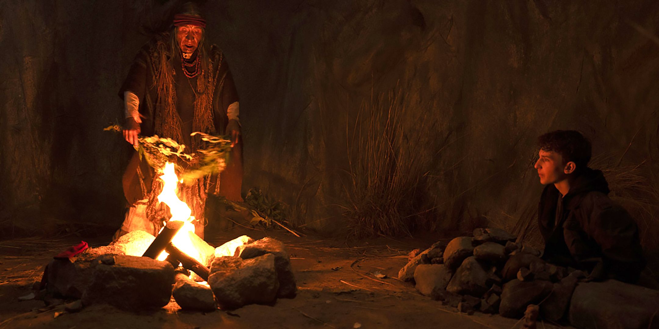 The shaman performing a fireside ritual while Elliot sits nearby in Shaman