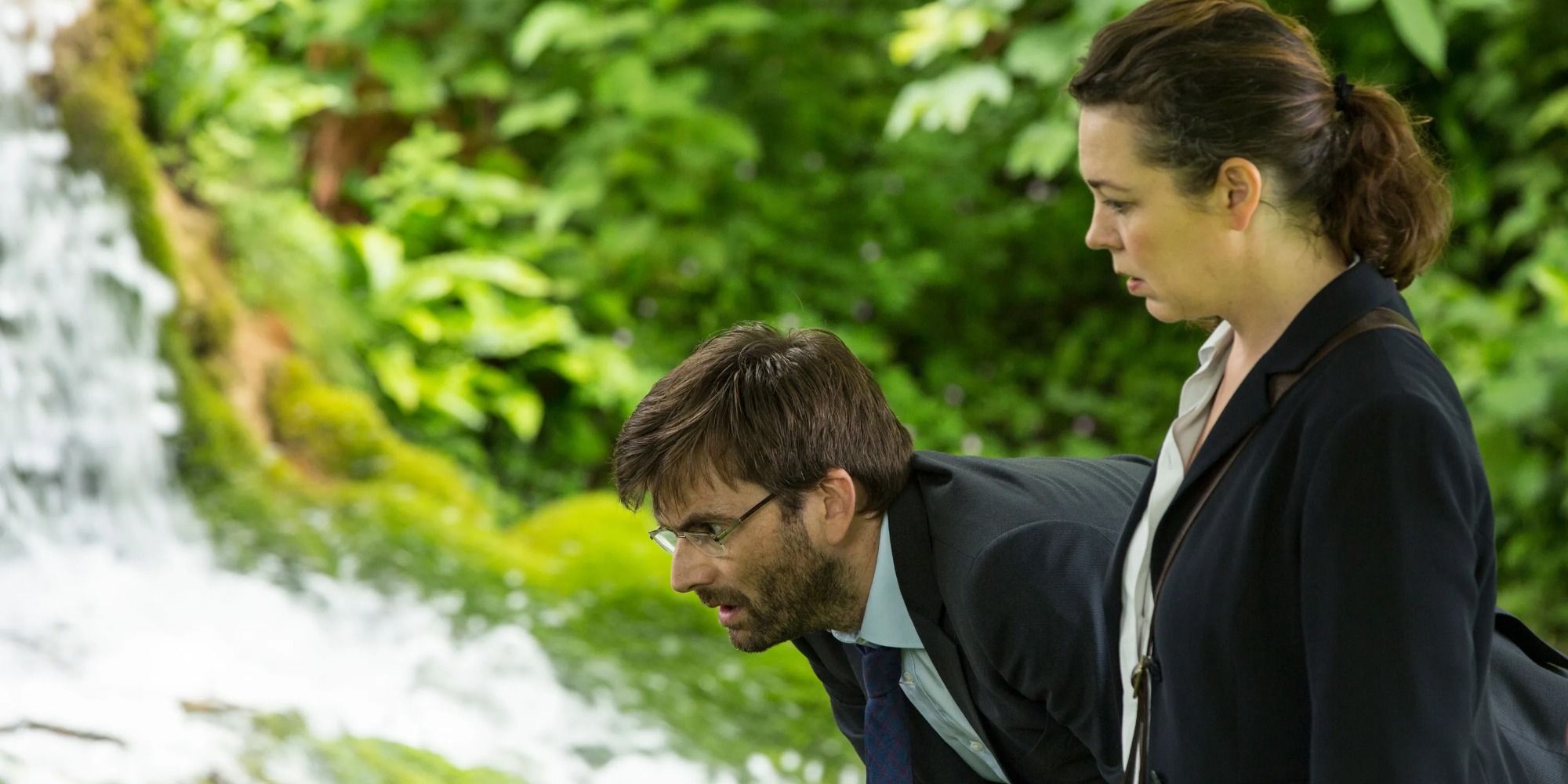 David Tennant como Alec Hardy e Olivia Colman como Ellie Miller em uma floresta, inclinando -se e franzindo a testa em alguma coisa, em Broadchurch.