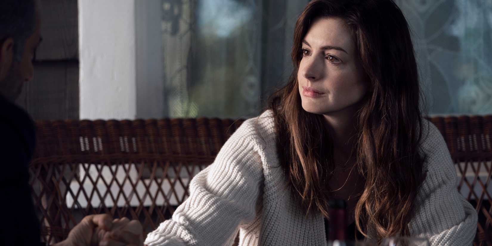 Anne Hathaway as Rebekah, holding someone's hand as she sits on a porch next to some wine glasses, in WeCrashed.