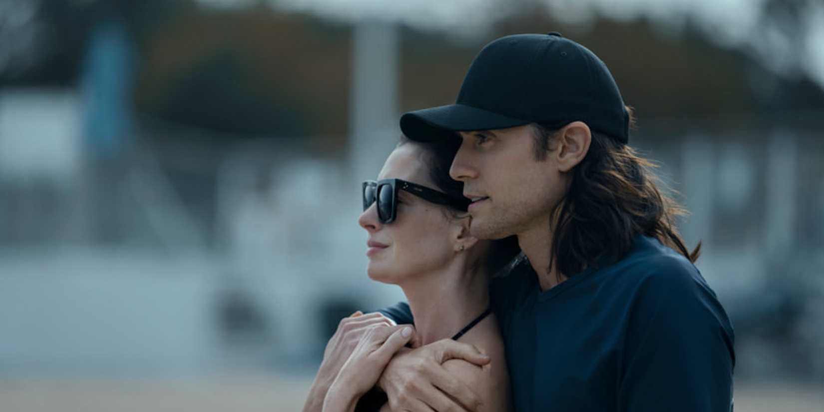Jared Leto like Adam wrapping her arms around Anne Hathaway like Rebekah while he stands outside, looking at something, in Wecrashed.