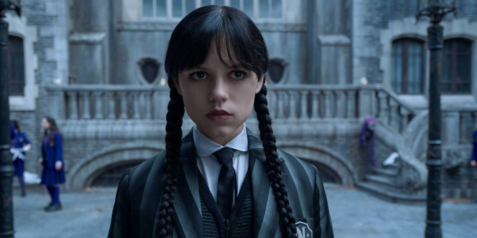 Jenna Ortega as Wednesday Addams looking deadpan while standing in front of Nevermore in Wednesday Season 2