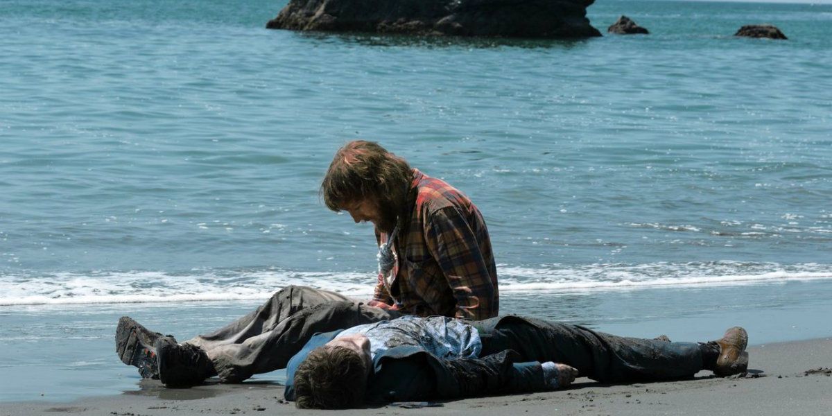 Manny lying on a beach while Hank kneels beside him in Swiss Army Man