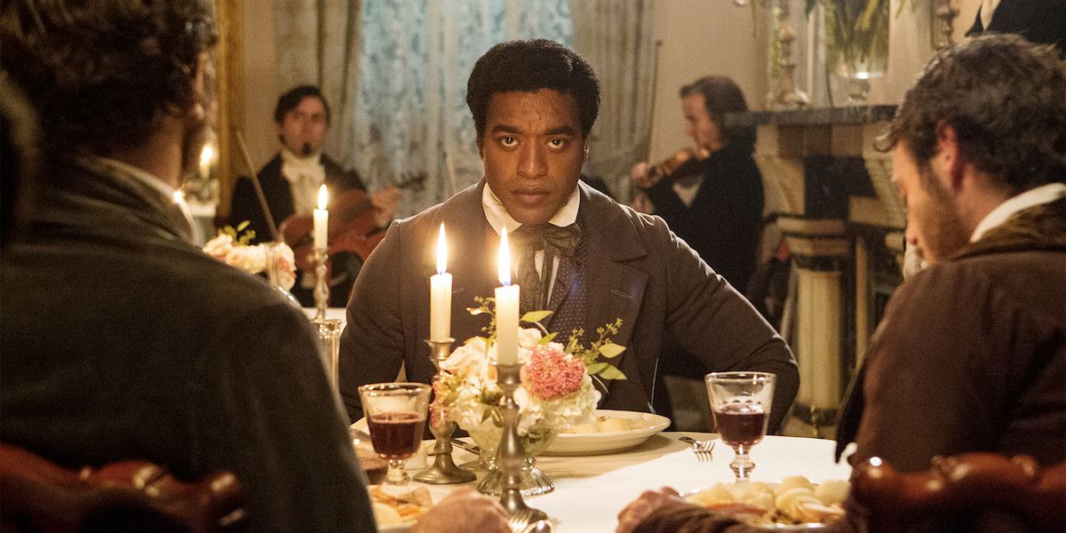 Chiwetel Ejiofor sits at a dining table in 12 years a slave, staring into the camera.