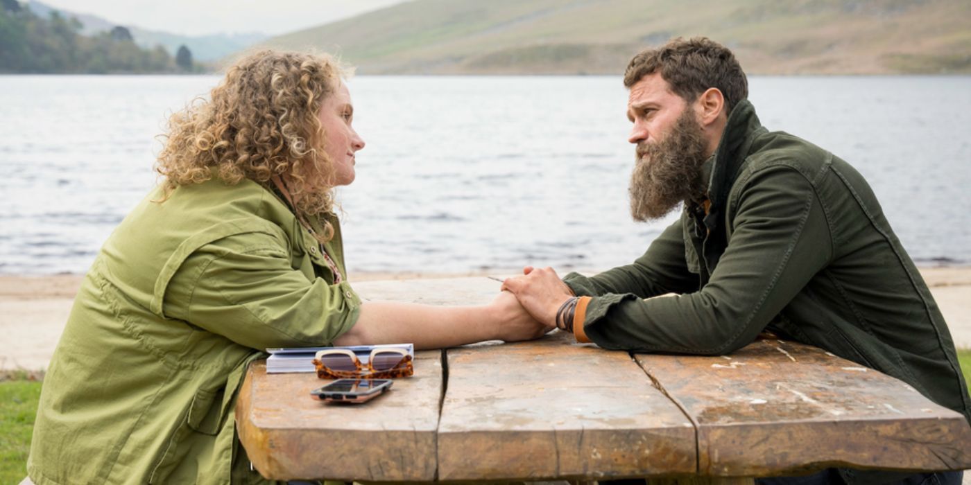 Danielle Macdonald as Helen Chambers and Jamie Dornan as Elliot hold hands at a picnic table in The Tourist season 2 - A Fargo Derivative, The Tourist Still Gives Genre Fans An Entertaining Ride